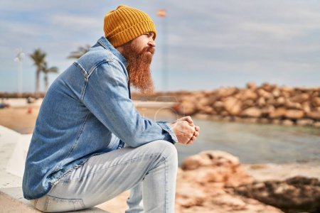 Foto de Young redhead man sitting on bench with serious expression at seaside - Imagen libre de derechos