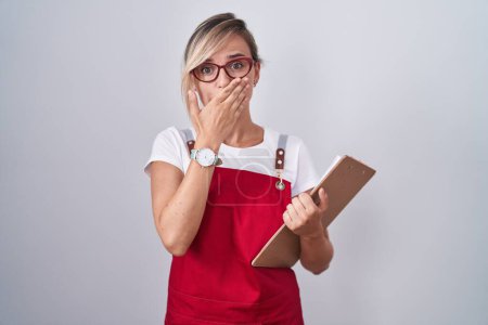 Photo for Young blonde woman wearing waiter uniform holding clipboard shocked covering mouth with hands for mistake. secret concept. - Royalty Free Image