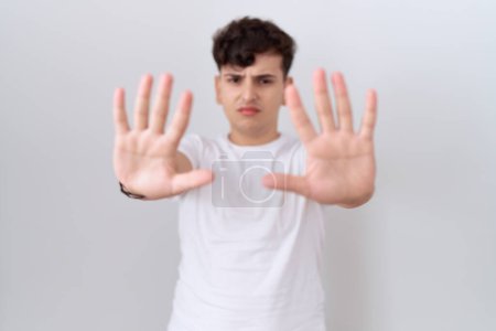 Téléchargez les photos : Jeune homme non binaire portant un t-shirt blanc décontracté faisant un geste d'arrêt avec les paumes des mains, expression de colère et de frustration - en image libre de droit