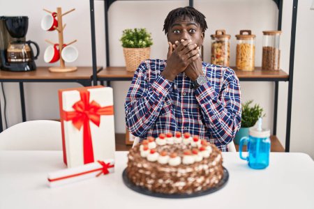 Téléchargez les photos : Homme africain avec dreadlocks célébrant anniversaire tenant grand gâteau au chocolat choqué couvrant la bouche avec les mains pour erreur. concept secret. - en image libre de droit