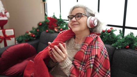 Téléchargez les photos : Femme du Moyen Âge aux cheveux gris écoutant de la musique boire du café assis par la décoration de Noël à la maison - en image libre de droit