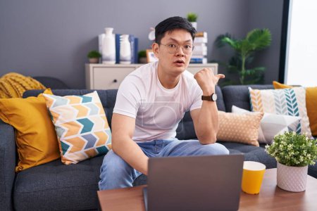 Foto de Joven asiático hombre usando portátil en casa sentado en el sofá sorprendido señalando con el dedo de la mano a un lado, boca abierta expresión asombrada. - Imagen libre de derechos