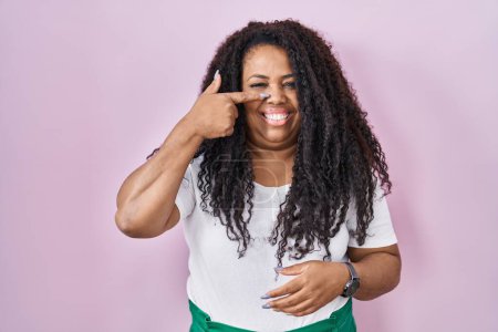 Téléchargez les photos : Plus taille hispanique femme debout sur fond rose pointant avec le doigt de la main vers le visage et le nez, souriant gai. concept de beauté - en image libre de droit