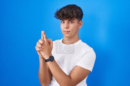 Photo for Hispanic teenager standing over blue background holding symbolic gun with hand gesture, playing killing shooting weapons, angry face - Royalty Free Image