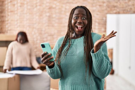 Téléchargez les photos : Femme africaine utilisant smartphone à la nouvelle maison célébrant la victoire avec sourire heureux et expression gagnante avec les mains levées - en image libre de droit