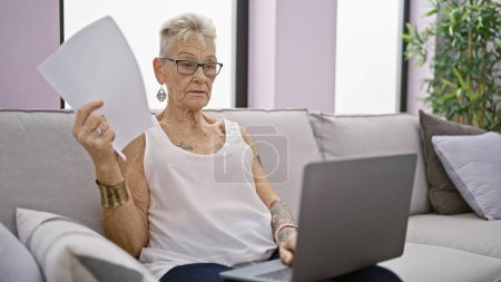 Téléchargez les photos : Femme âgée aux cheveux gris concentrée, profondément concentrée, utilisant son ordinateur portable et tenant des documents tout en étant confortablement assise à la maison sur son canapé du salon. - en image libre de droit