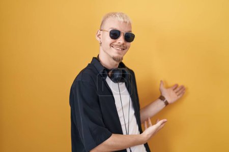 Photo for Young caucasian man wearing sunglasses standing over yellow background inviting to enter smiling natural with open hand - Royalty Free Image