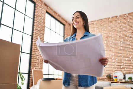 Photo for Young beautiful hispanic woman architect smiling confident reading house plans at new home - Royalty Free Image