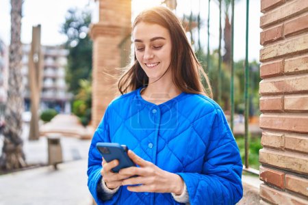 Téléchargez les photos : Jeune femme caucasienne souriant confiant en utilisant un smartphone à la rue - en image libre de droit