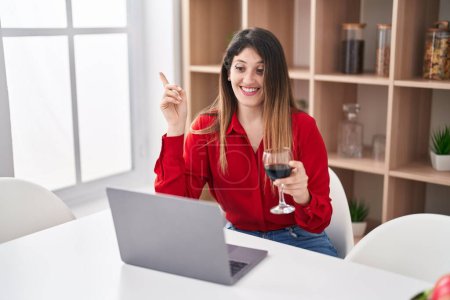 Photo for Young brunette woman doing video call drinking red wine smiling happy pointing with hand and finger to the side - Royalty Free Image
