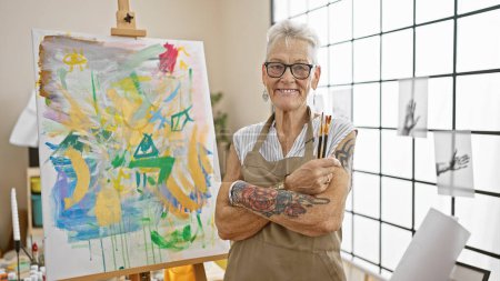 Foto de Sonriente artista senior de cabello gris de pie con un gesto relajado de brazos cruzados, disfrutando de su pasatiempo entre el lienzo y los pinceles en su estudio de arte. - Imagen libre de derechos