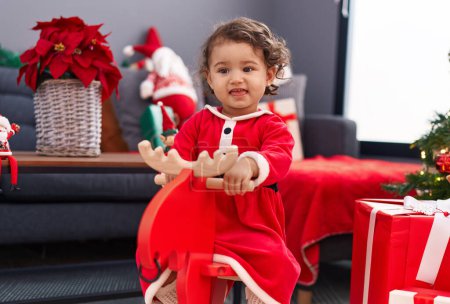 Photo for Adorable hispanic toddler playing with reindeer rocking by christmas tree at home - Royalty Free Image