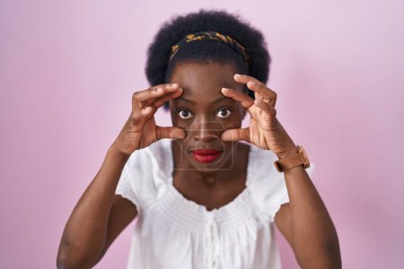 Foto de Mujer africana con el pelo rizado de pie sobre fondo rosa tratando de abrir los ojos con los dedos, somnoliento y cansado para la fatiga de la mañana - Imagen libre de derechos