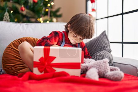 Téléchargez les photos : Adorable tout-petit déballer cadeau de Noël assis sur le canapé à la maison - en image libre de droit
