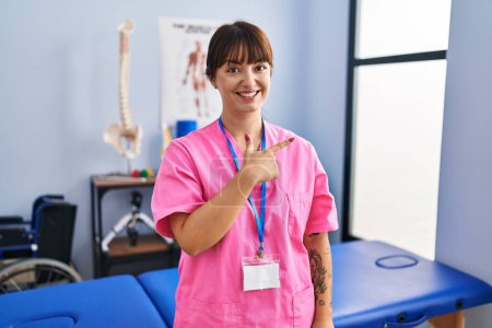 Photo for Young brunette woman working at rehabilitation clinic cheerful with a smile of face pointing with hand and finger up to the side with happy and natural expression on face - Royalty Free Image