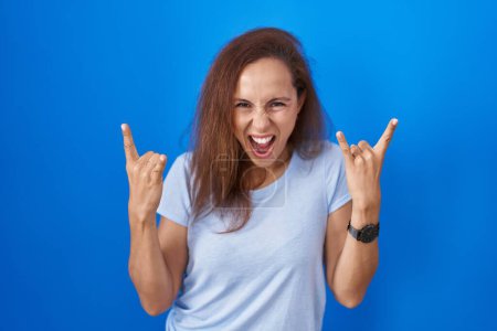 Photo for Brunette woman standing over blue background shouting with crazy expression doing rock symbol with hands up. music star. heavy music concept. - Royalty Free Image