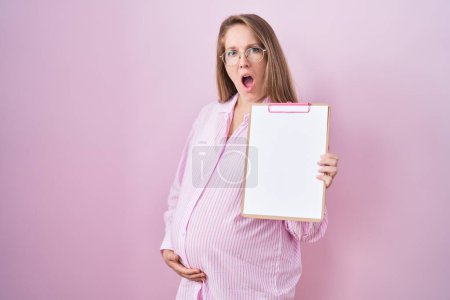 Téléchargez les photos : Jeune femme enceinte tenant presse-papiers dans le visage de choc, l'air sceptique et sarcastique, surpris avec la bouche ouverte - en image libre de droit