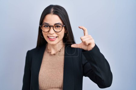 Téléchargez les photos : Jeune femme brune debout sur fond bleu geste souriant et confiant avec la main faisant signe de petite taille avec les doigts regardant et la caméra. concept de mesure. - en image libre de droit