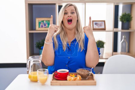 Téléchargez les photos : Caucasienne plus la taille femme manger le petit déjeuner à la maison étonné et surpris de regarder vers le haut et pointant avec les doigts et les bras levés. - en image libre de droit