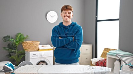Photo for Young hispanic man standing with arms crossed gesture by ironing board at laundry room - Royalty Free Image