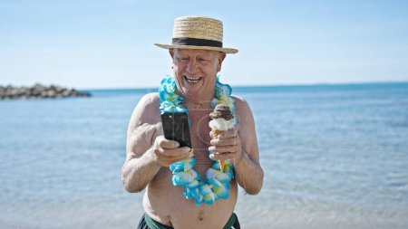 Téléchargez les photos : Homme âgé aux cheveux gris touriste tenant de la crème glacée en utilisant un smartphone au bord de la mer - en image libre de droit
