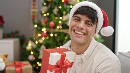 Téléchargez les photos : Jeune homme hispanique souriant confiant tenant cadeau de Noël à la maison - en image libre de droit