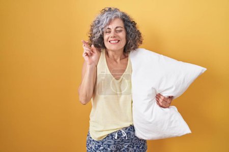 Téléchargez les photos : Femme d'âge moyen avec les cheveux gris portant un pyjama étreignant l'oreiller geste doigt croisé souriant avec espoir et les yeux fermés. chance et concept superstitieux. - en image libre de droit