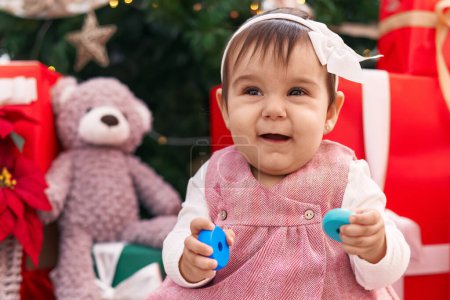 Foto de Adorable bebé hispano sosteniendo aros de juguete sentado en el suelo por el árbol de Navidad en casa - Imagen libre de derechos