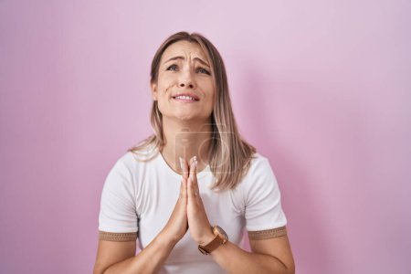 Photo for Blonde caucasian woman standing over pink background begging and praying with hands together with hope expression on face very emotional and worried. begging. - Royalty Free Image
