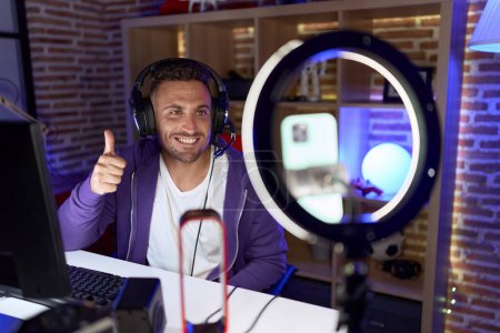 Foto de Hombre hispano con barba jugando videojuegos grabando con teléfono inteligente sonriendo feliz y positivo, pulgar hacia arriba haciendo excelente y signo de aprobación - Imagen libre de derechos