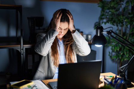 Téléchargez les photos : Jeune femme brune travaillant au bureau la nuit souffrant de maux de tête désespérés et stressés à cause de la douleur et de la migraine. mains sur la tête. - en image libre de droit