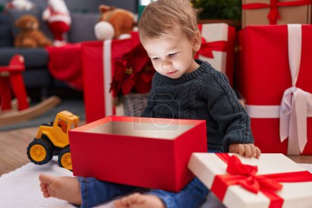 Foto de Adorable niño rubio desempacar regalo de Navidad sentado en el suelo en casa - Imagen libre de derechos