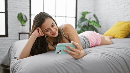 Téléchargez les photos : Joyeux jeune femme hispanique couchée au lit, en pyjama, ayant un appel vidéo heureux dans la chambre confortable de son appartement - en image libre de droit