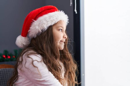 Photo for Adorable hispanic girl wearing christmas hat looking through the window at home - Royalty Free Image