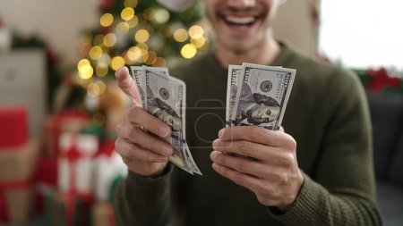 Foto de Joven hombre hispano contando dólares sentado en el sofá por el árbol de Navidad en casa - Imagen libre de derechos