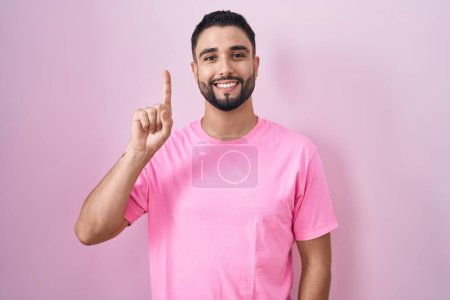 Téléchargez les photos : Jeune homme hispanique debout sur fond rose montrant et pointant vers le haut avec le doigt numéro un tout en souriant confiant et heureux. - en image libre de droit