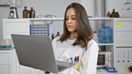Téléchargez les photos : Jeune femme hispanique concentrée, une belle scientifique au travail, utilisant un ordinateur portable et un microscope pour la recherche médicale en laboratoire, affichant une expression détendue, mais déterminée - en image libre de droit