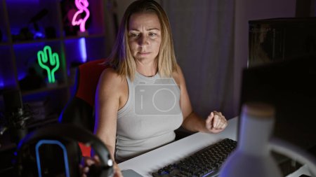 Photo for Tense night, attractive young blonde woman streamer, headphones clutched, eyes focused online - inside the dark of a home gaming room, engrossed in the futuristic world of cyber gaming. - Royalty Free Image
