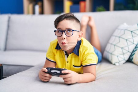 Foto de Joven niño hispano jugando videojuego sosteniendo controlador en el sofá haciendo cara de pez con boca y ojos entrecerrados, loco y cómico. - Imagen libre de derechos