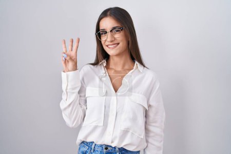 Téléchargez les photos : Jeune femme brune portant des lunettes montrant et pointant vers le haut avec les doigts numéro trois tout en souriant confiant et heureux. - en image libre de droit