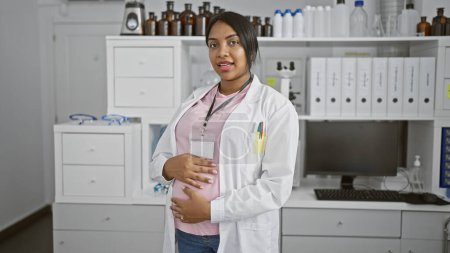Foto de Esperando científico, joven, mujer embarazada, sonriendo mientras toca su vientre, en medio de la investigación médica en el laboratorio - Imagen libre de derechos
