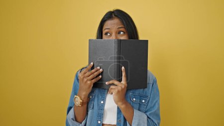 Photo for African american woman covering mouth with book over isolated yellow background - Royalty Free Image