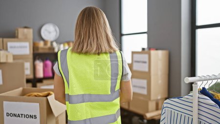Photo for Attractive young blonde volunteer in reflective vest standing with her back to charity center, working at indoor warehouse, promoting community service and altruism. - Royalty Free Image