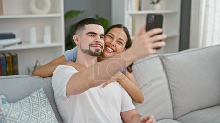Téléchargez les photos : Beau couple faisant plaisir souvenirs selfie sur le canapé, souriant en toute confiance et exprimant l'amour à leur maison confortable - en image libre de droit