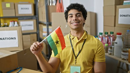 Téléchargez les photos : Jeune homme latin volontaire tenant drapeau ghana souriant au centre de charité - en image libre de droit
