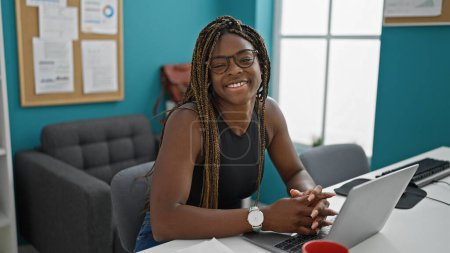 Téléchargez les photos : Afro-américaine femme d'affaires en utilisant ordinateur portable souriant au bureau - en image libre de droit
