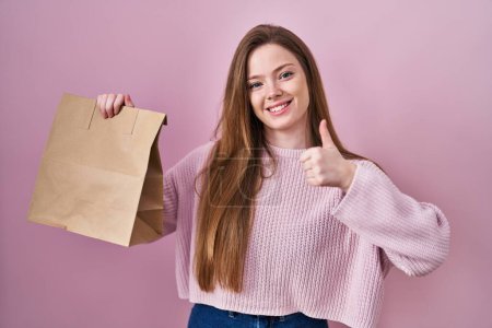 Foto de Joven mujer caucásica sosteniendo bolsa de compras y tarjeta de crédito sonriendo feliz y positivo, pulgar hacia arriba haciendo excelente y signo de aprobación - Imagen libre de derechos