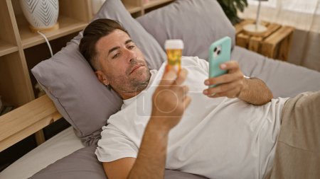 Téléchargez les photos : Troublé jeune homme hispanique couché malade en pyjama sur son lit, regardant anxieusement son smartphone tout en saisissant une bouteille de pilules dans sa chambre. - en image libre de droit