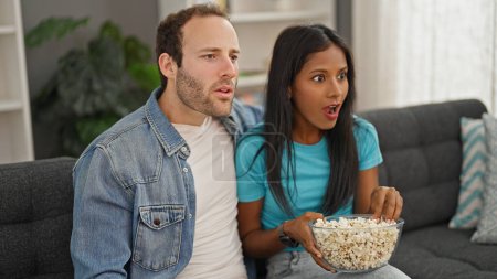 Photo for Beautiful couple watching tv eating popcorn surprised at home - Royalty Free Image