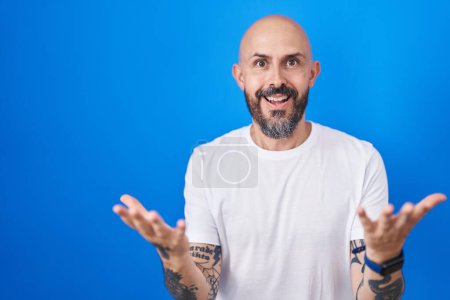 Téléchargez les photos : Homme hispanique avec des tatouages debout sur fond bleu souriant gai à bras ouverts comme accueil amical, salutations positives et confiantes - en image libre de droit